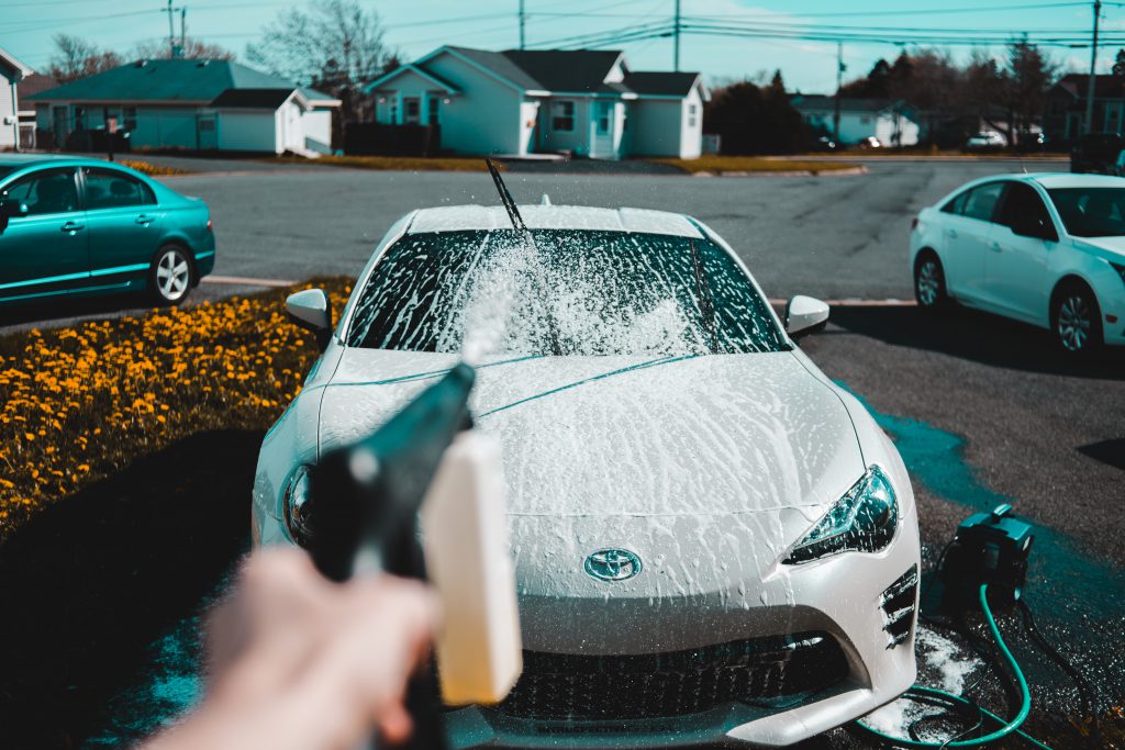 the photographer's left hand holds a hose with an additional attachment containing soap, spraying a white car parked on a driveway. a road curves around in the background with white houses lining the street and two cars just visible either side of the white car being cleaned.